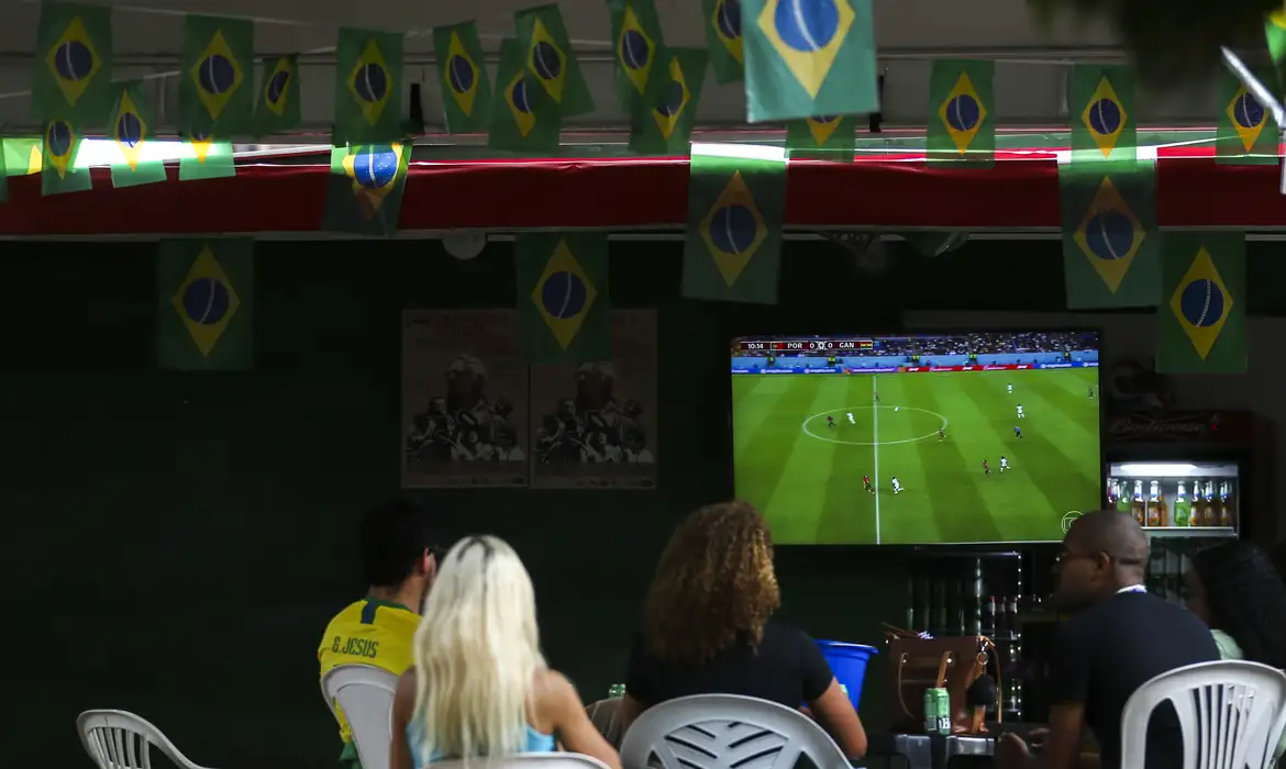 Brasil nas oitavas da Copa pode jogar em horário comercial; veja os impactos 6 Pessoas assistem partida de futebol da Seleção Brasileira em bar decorado com bandeiras do Brasil