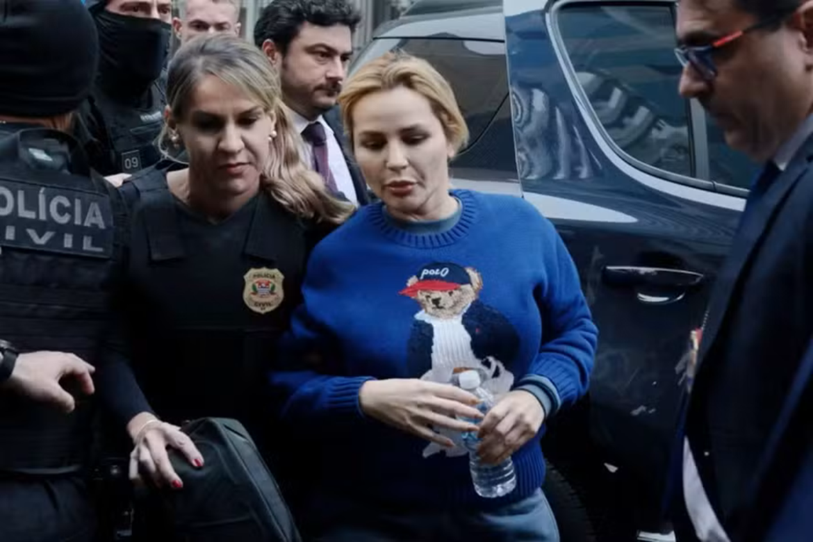 Mulher de cabelos loiros com suéter azul é escoltada por policiais civis, segurando garrafa de água, próximo a viatura