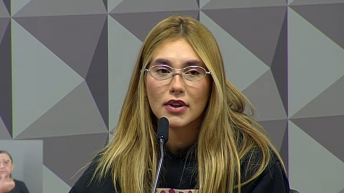 Mulher loira de óculos e blusa preta fala ao microfone em ambiente com painel geométrico cinza e azul