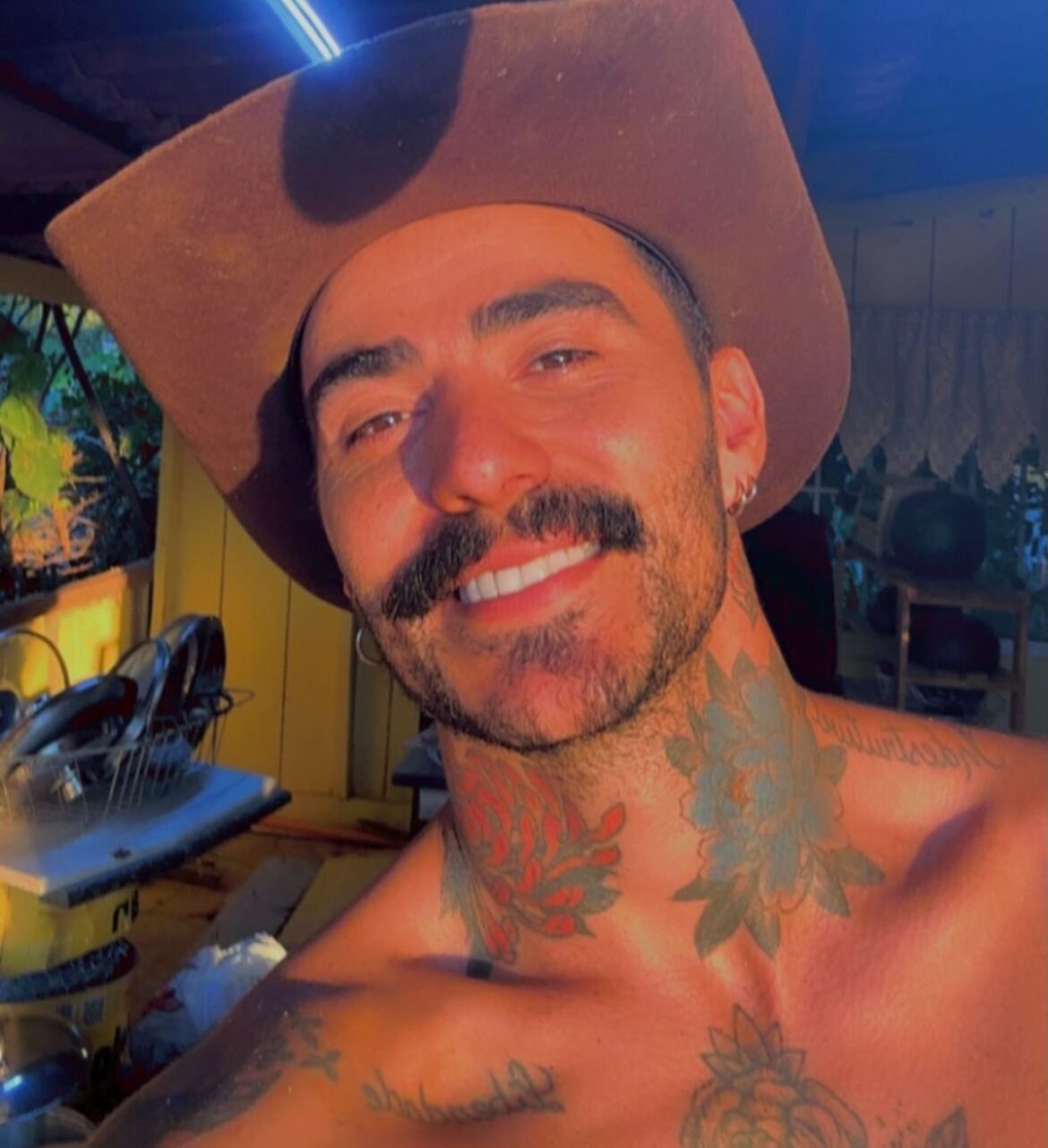 Homem sorri em selfie usando chapéu de couro com tatuagens visíveis no pescoço
