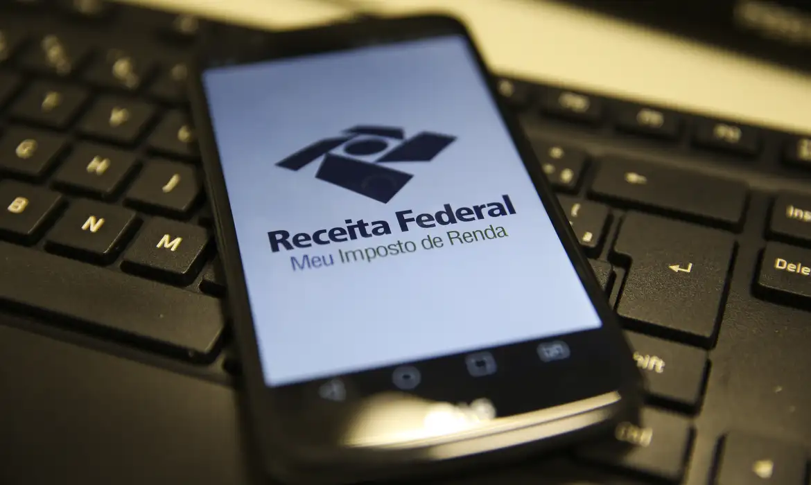 IRPF 2026: Receita libera consulta ao 3º lote a mais de 2,7 milhões de contribuintes nesta sexta (24) 6 Smartphone exibe tela da Receita Federal Meu Imposto de Renda sobre teclado de computador preto