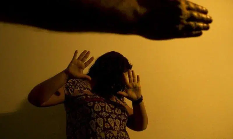 Feminicídios batem recorde em 2025: 1 em cada 9 vítimas tinha medida protetiva ativa, aponta Anuário 6 Mulher levanta as mãos tentando se proteger de um braço levantado contra ela em fundo amarelo escuro